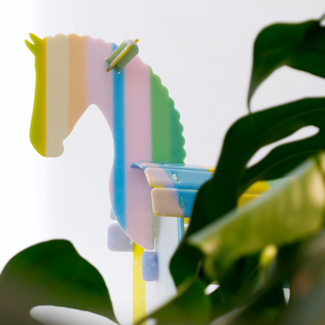 Precious Things; Rocking Horse — vertical detail of the sculpture's head in pastel stripes, seen through leaves
